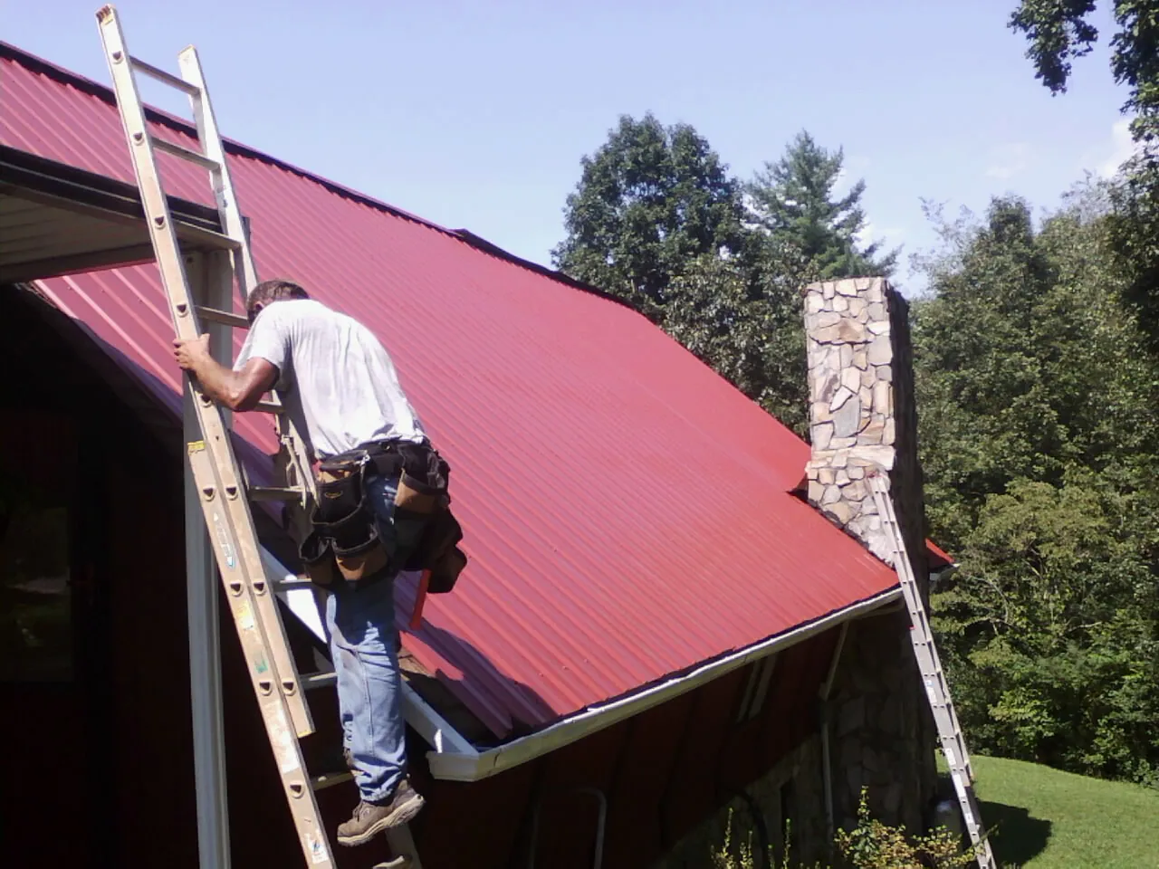 Roofer on a ladder beside a red metal roof and stone chimney — Soffit & Fascia Repair by Roofing Brothers in Silverstreet